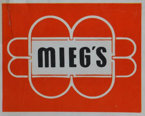 emblem aus Längs- und Quer-Ellipsen, darin "MIEG'S" in Rot, auch MIEG's in schwarz auf weißem Rechteck in Ellipsen-Mitte
