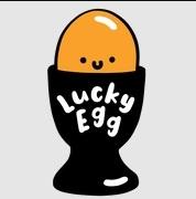 schwarzer Eierbecher, darin dunkeloranges Ei mit punkt-.-Punkt-Strich Gesicht, Lucky Egg in zwei Zeilen in oberer Hälfte des Eierbechers